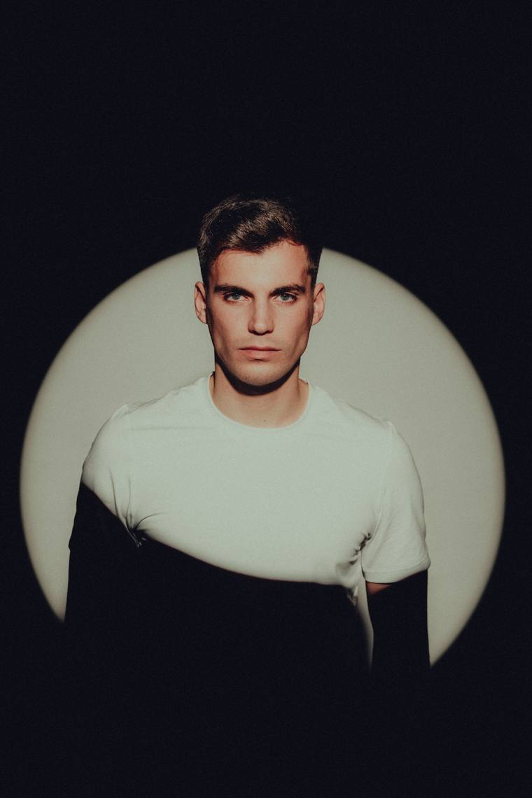 Bence Laska in a white T-shirt, standing in a round spotlight on a dark background